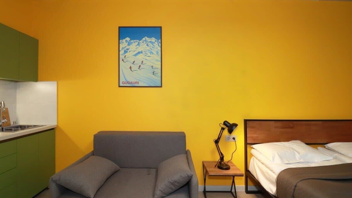 Ein Hotelzimmer mit Bett, Sofa, Nachttisch mit Lampe und einer Küchenzeile mit Spüle und Schränken.