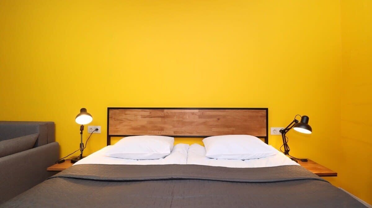 A bed with a wooden headboard, two bedside lamps, and a grey sofa in a room with yellow walls.