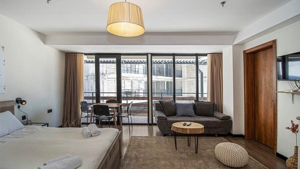 A hotel room with a bed, a sofa, a small table, a chair, a TV, and a large window with a view of buildings. - Cozy 1BR Ski Resort Apt in Gudauri - By Wehost (Gudauri)