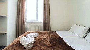 Une chambre d’hôtel avec un lit, une fenêtre avec des rideaux, un radiateur et une étagère sur laquelle se trouve un petit objet.