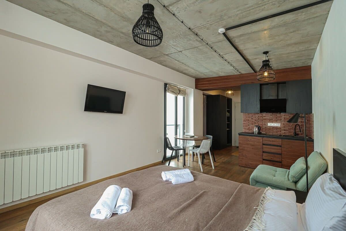 A modern hotel room with a bed, a dining area, and a kitchenette.