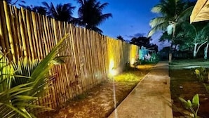 Une vue nocturne d’un sentier bordé d’une clôture en bambou et de palmiers, menant à un bâtiment doté d’un toit de chaume.