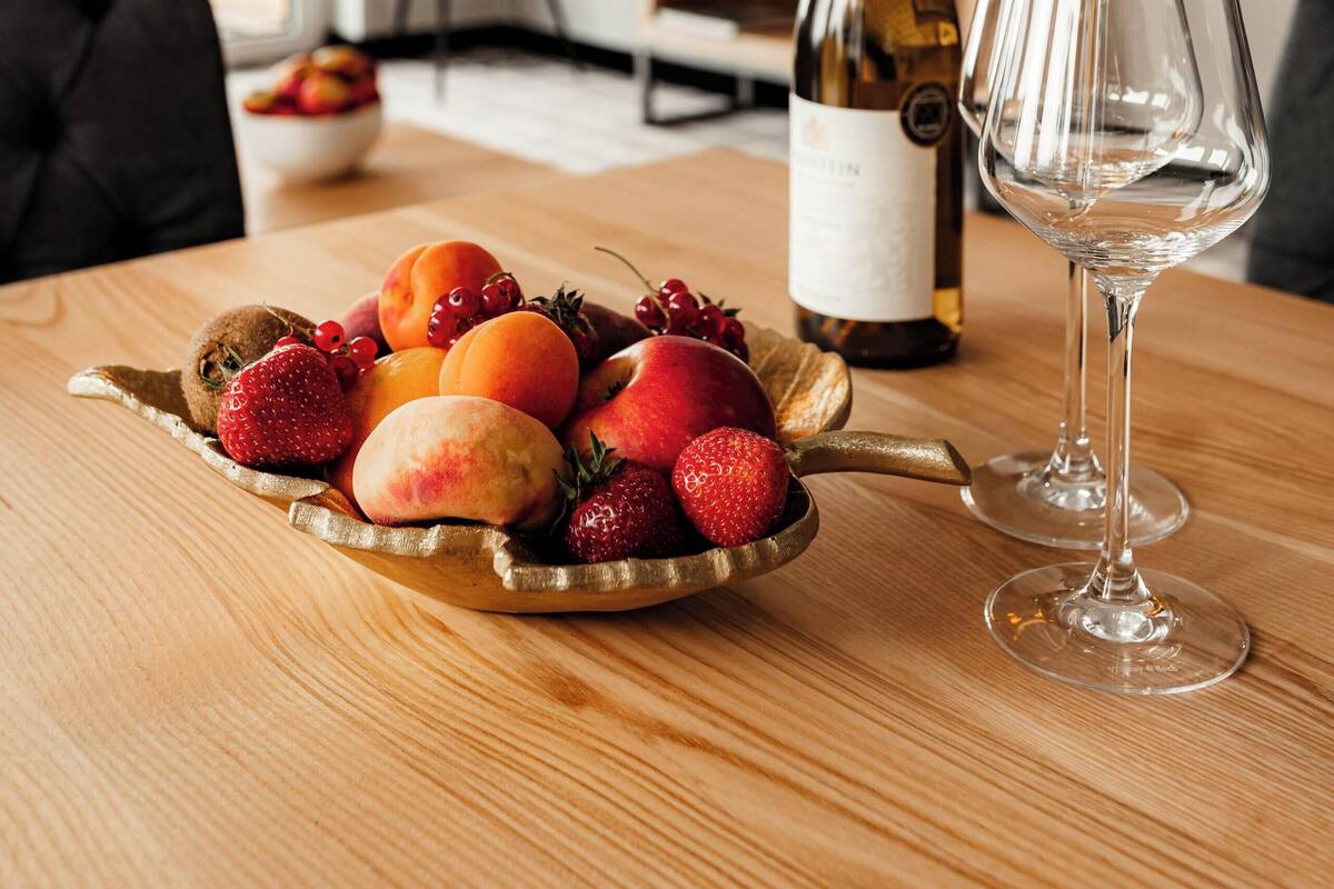 A wooden table with a fruit bowl, a wine bottle, and a wine glass in a room with a TV and a lamp in the background.