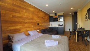 Una habitación de hotel con pared de madera, una cama con almohadas, una mesita de noche, una pequeña cocina con microondas y una mesa de comedor con sillas.