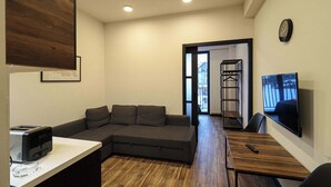 A modern living room with a sectional sofa, a dining table, a flat-screen TV, and a kitchen area with a toaster. - Brand New Ski 1BR Apt In New Gudauri - By Wehost (Gudauri)