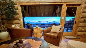 A cozy log cabin interior with leather furniture, a large window offering a mountain view, and a rustic wooden floor. - Sunrise Ridge – Lake & Mountain View Luxury (Blue Ridge)