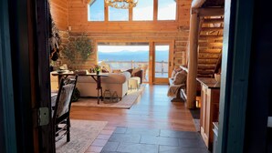 A log cabin interior with a wooden floor, a chandelier, a sofa, a dining table, and a view of the mountains through large windows. - Sunrise Ridge – Lake & Mountain View Luxury (Blue Ridge)