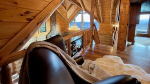 A cozy log cabin interior with a stone fireplace, wooden beams, and a large window offering a view of the mountains. - Sunrise Ridge – Lake & Mountain View Luxury (Blue Ridge)