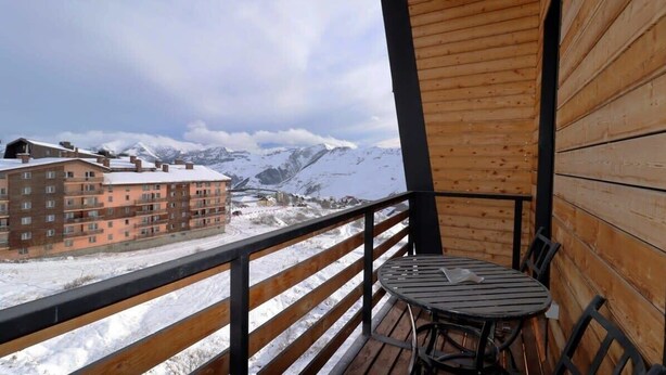A wooden balcony with a table and chairs, overlooking a snowy mountain landscape and buildings. - Modern Mountain Retreat in Gudauri – By Wehost (Gudauri)