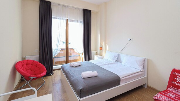 A bedroom with a bed, a red chair, a white bedside table, a lamp, and a window with curtains. - Charming New Gudauri Studio - by Wehost (Gudauri)