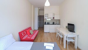 A compact room with a bed, a red sofa with patterned cushions, a dining table with chairs, a small kitchen area, and a TV. - Charming New Gudauri Studio - by Wehost (Gudauri)