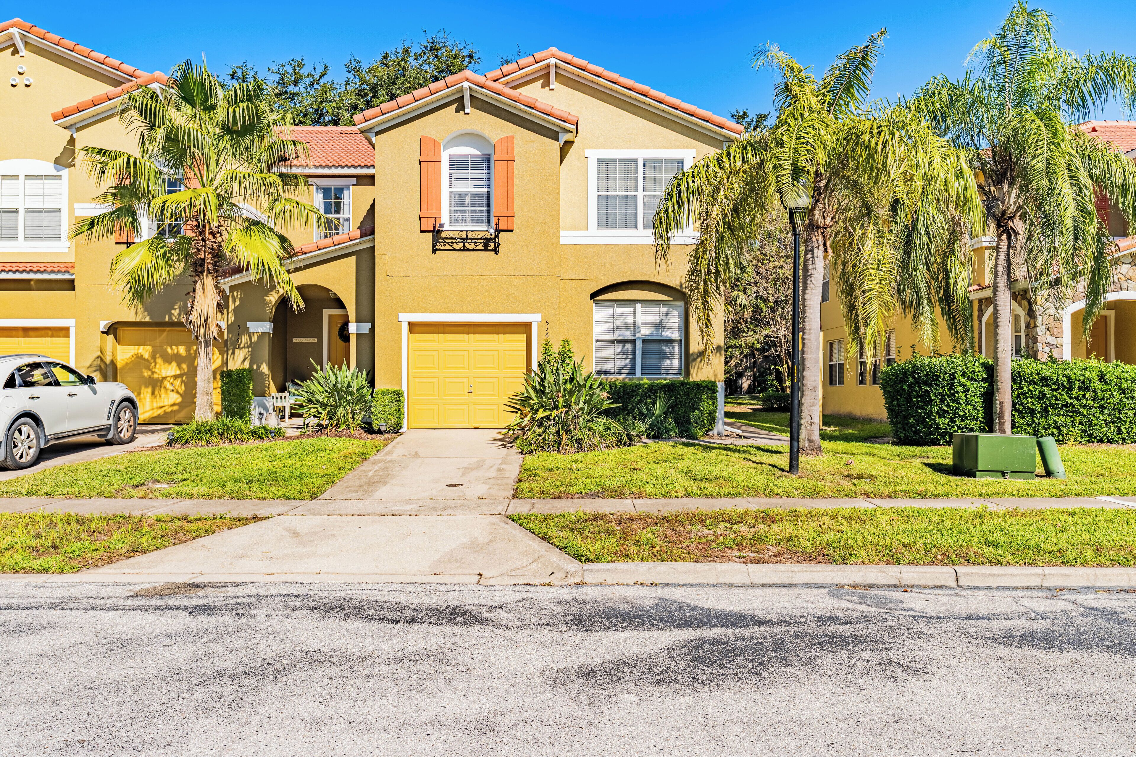 A two-story yellow house with a red-tiled roof, a garage, and a driveway leading to the entrance.