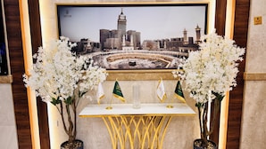A reception area with a framed cityscape photograph, two potted plants with white flowers, and a table with two flags. - alryada grand (Makkah)