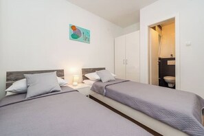Un dormitorio con dos camas, una mesita de noche con lámpara y un baño visible a través de una puerta abierta.