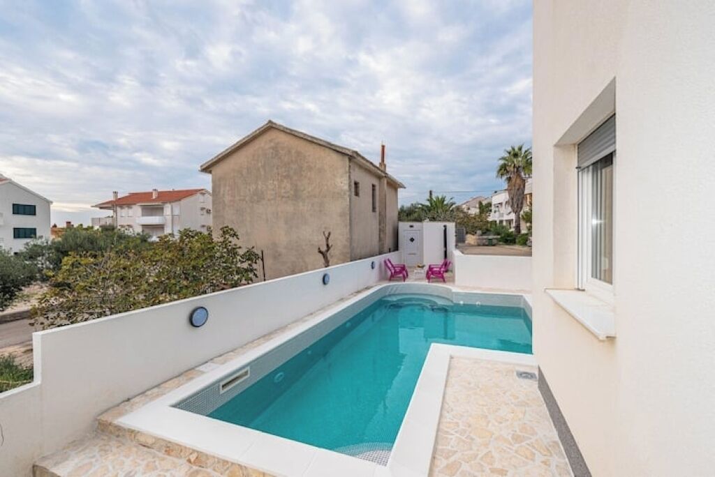 A swimming pool with a stone deck, a small building in the background, and a clear sky.