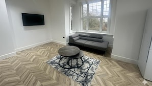 Een moderne woonkamer met een visgraatvloer, een grijze bank, een ronde salontafel en een groot raam dat voor natuurlijk licht zorgt.