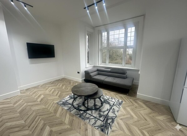 A modern living room with a herringbone floor, a grey sofa, a round coffee table, and a large window providing natural light. - Dippen Hall Apartment - Minutes From London Gatwick (Lingfield)