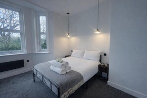 A neatly made bed with rolled towels, a bedside table, and a nightstand. There are two windows with white frames and a view of trees outside. - Dippen Hall Apartment - Minutes From London Gatwick (Lingfield)