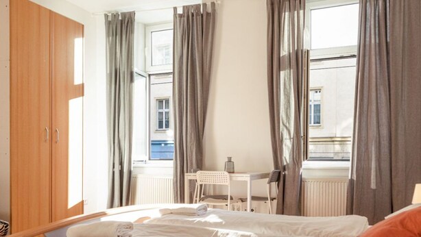 Ein Schlafzimmer mit einem Bett, einem Schreibtisch, einem Stuhl, einem Holzschrank und einem Fenster mit Vorhängen.