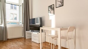 Ein Zimmer mit Fernseher, einem kleinen weißen Tisch, einem Stuhl, einem Fenster mit Vorhängen und zwei gerahmten Bildern an der Wand.