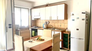 Eine kleine Küche mit weißen Schränken, einem Herd, einer Spüle und einem Kühlschrank. Ein Esstisch mit Stühlen und ein kleines Fenster sorgen für natürliches Licht.