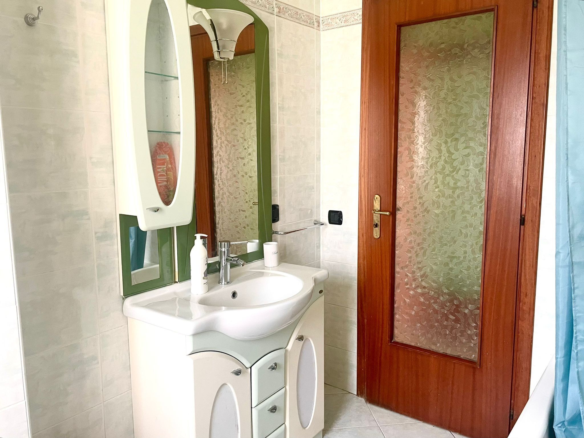 Une salle de bain avec un lavabo blanc, un miroir, un miroir encadré de vert, une porte en bois avec un panneau en verre dépoli et des toilettes.