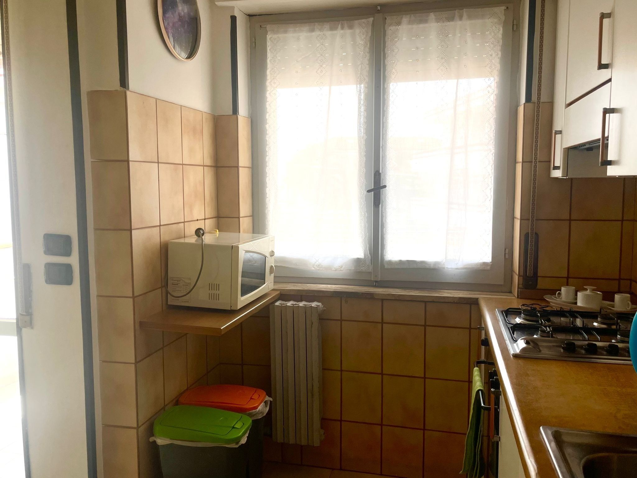 Uma cozinha com micro-ondas numa prateleira, janela com cortinas, fogão e paredes revestidas de azulejos.
