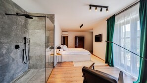 A modern hotel room with a glass shower enclosure, a bed, a desk, and a large window with curtains.