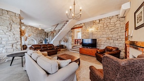 A living room with stone walls, a staircase, a chandelier, leather sofas, a TV, and a fireplace.