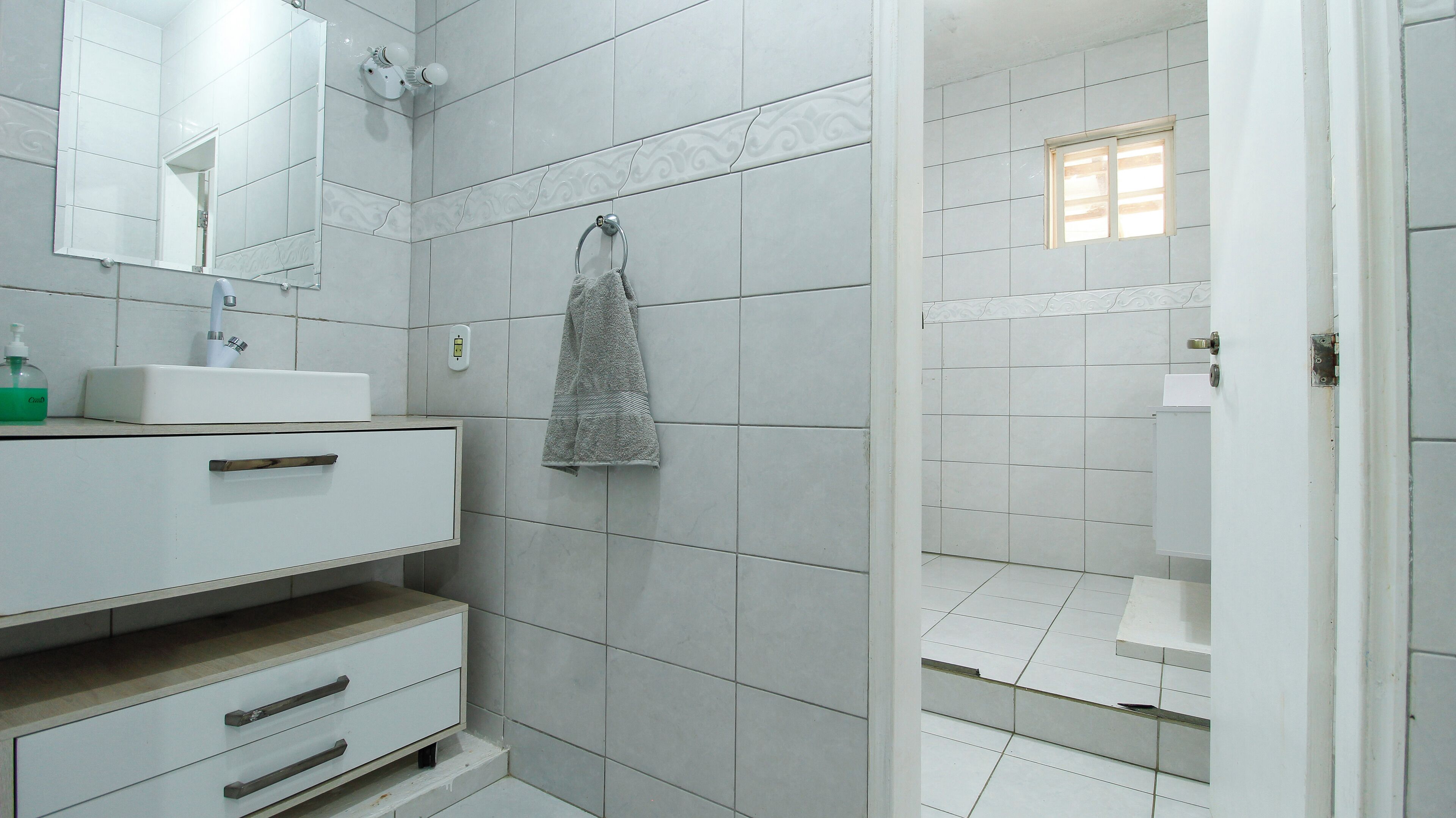 Un baño con azulejos blancos, un lavamanos, un espejo, toallero y zona de ducha.