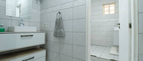 Un baño con azulejos blancos, un lavamanos, un espejo, toallero y zona de ducha.