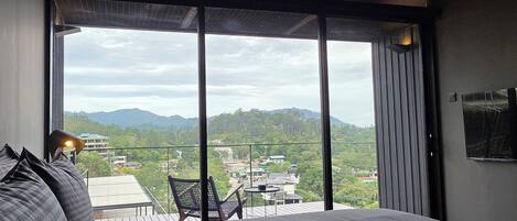 A modern hotel room with a large bed, a small table, and a chair. The room has a large window offering a view of a cityscape and mountains.