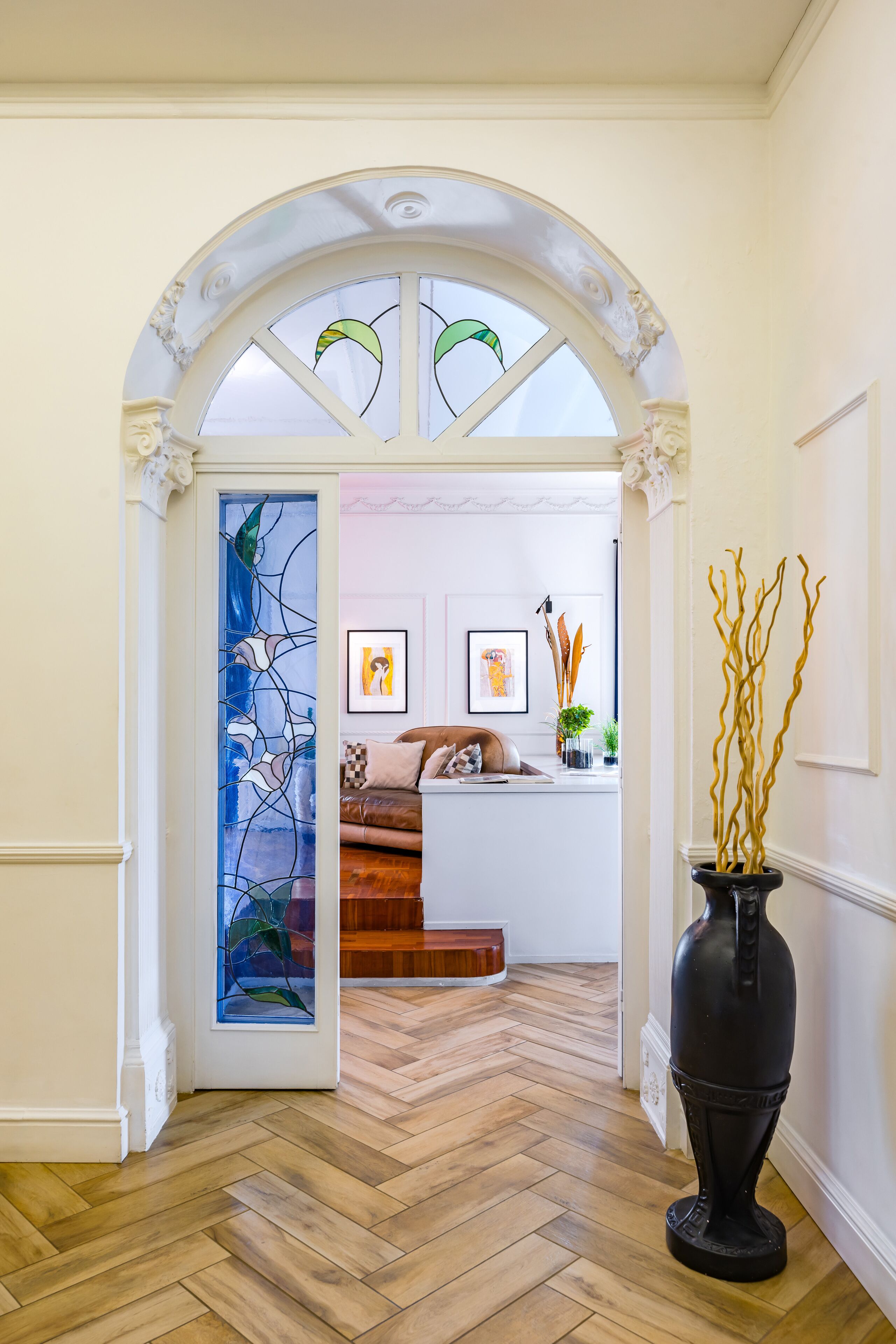A hallway with a wooden herringbone floor, a black vase with golden branches, and a stained glass door with abstract designs.