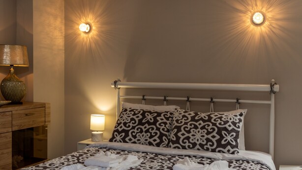 Una cama bien hecha con ropa de cama estampada, una lámpara de noche y una lámpara fijada a la pared.