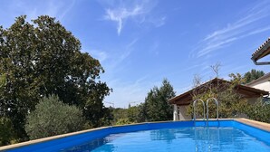 Una piscina cristallina con bordi in legno, circondata da alberi e un edificio con tetto spiovente.