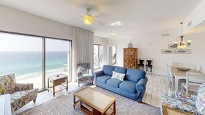 Ein geräumiges Wohnzimmer mit großem Fenster mit Meerblick, ein blaues Sofa, ein gemustertes Sessel und eine Essecke mit einer Chandelier.