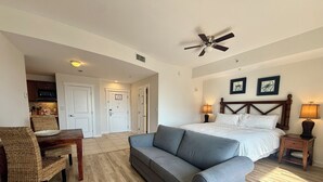 Habitación de hotel con una cama, un sofá gris, un escritorio de madera, sillas de mimbre y un ventilador de techo.