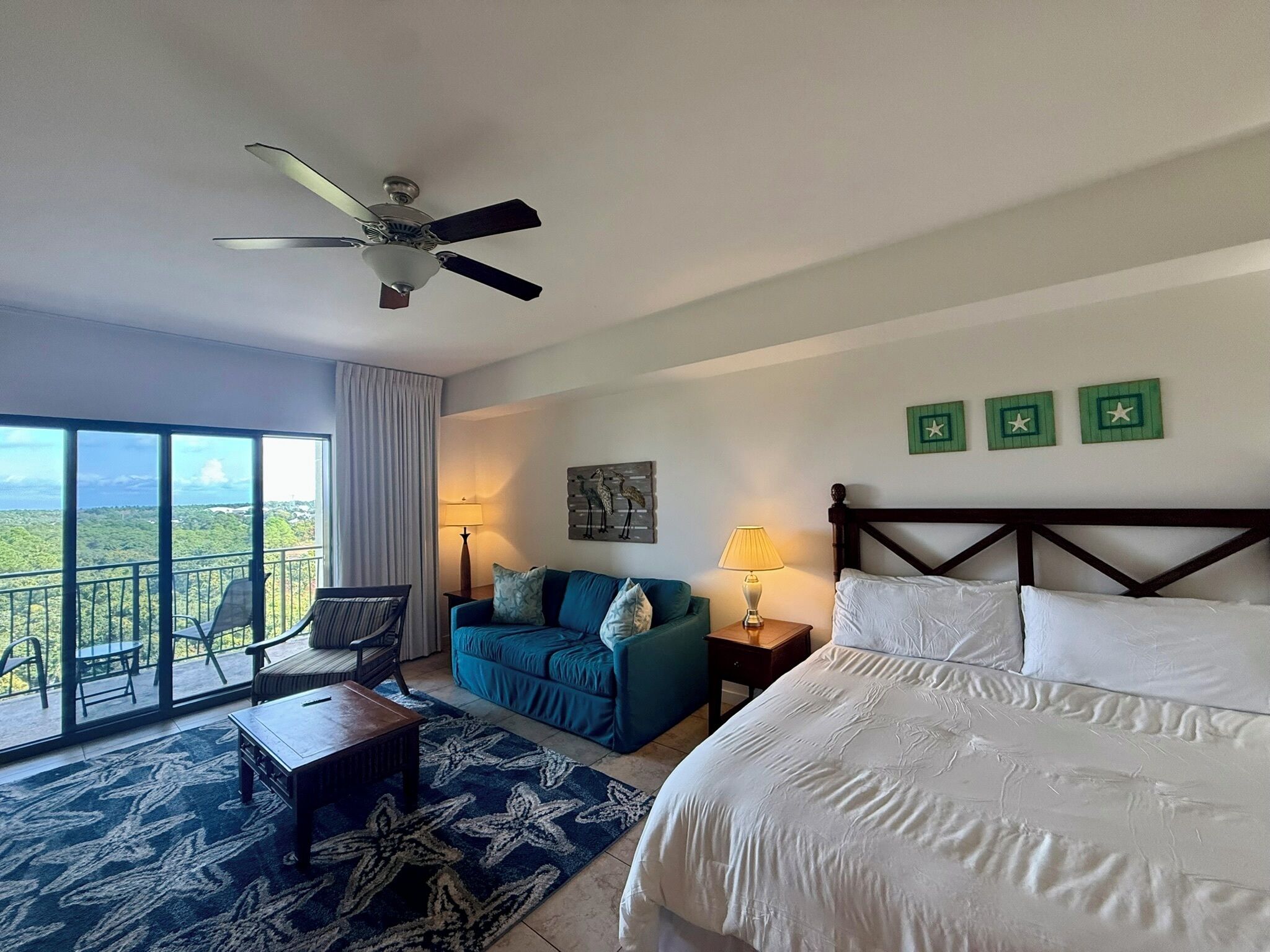 A hotel room with a bed, a sofa, a chair, a table, a lamp, a ceiling fan, and a view of the landscape.