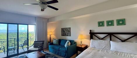 A hotel room with a bed, a sofa, a chair, a table, a lamp, a ceiling fan, and a view of the landscape.