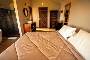 A bedroom with a bed, a wooden wardrobe, a bathroom with a glass shower, and a door leading to another room.
