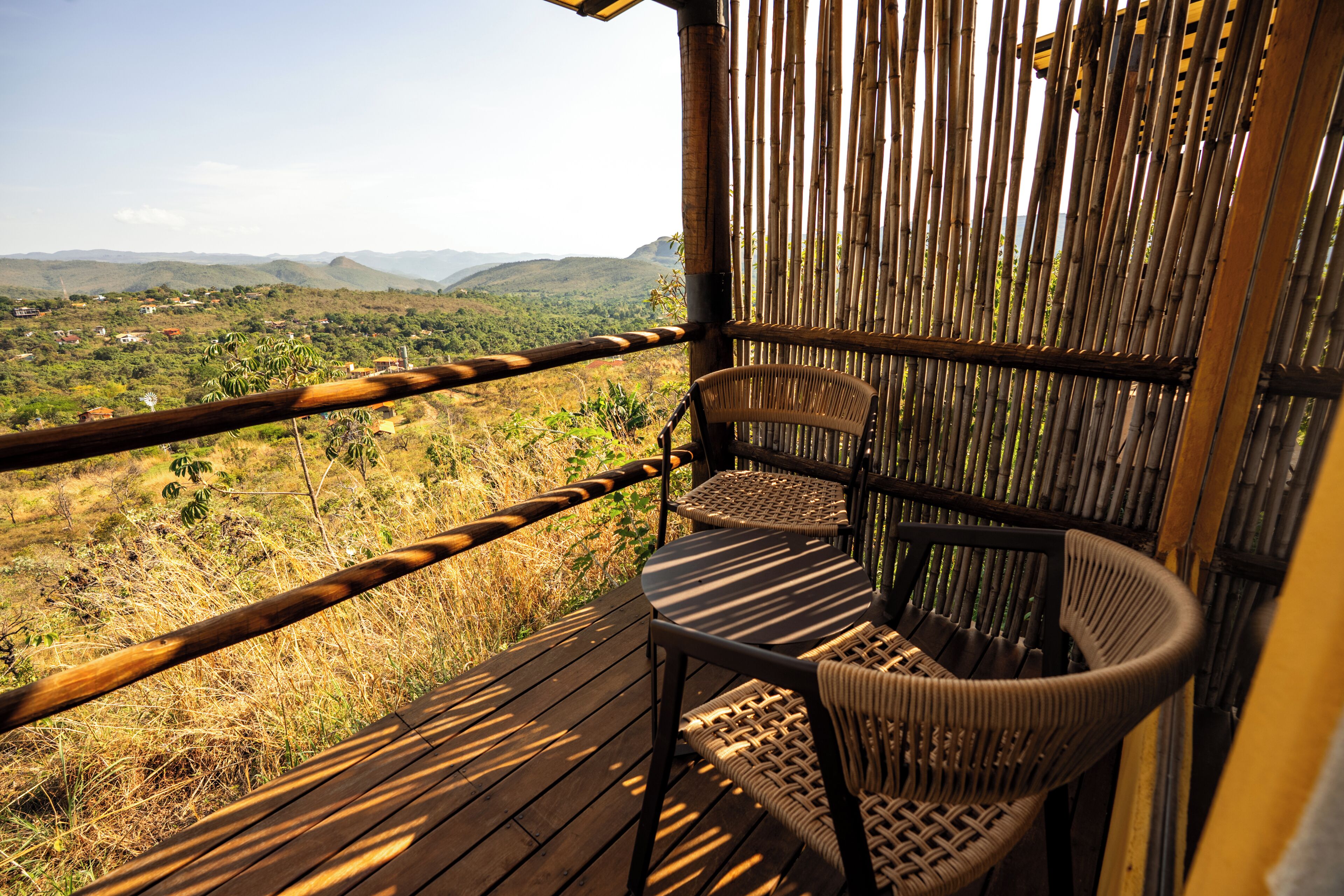 Un balcon doté d’un plancher en bois, d’une chaise en osier et d’une clôture en bambou, offrant une vue sur un paysage vallonné et verdoyant.