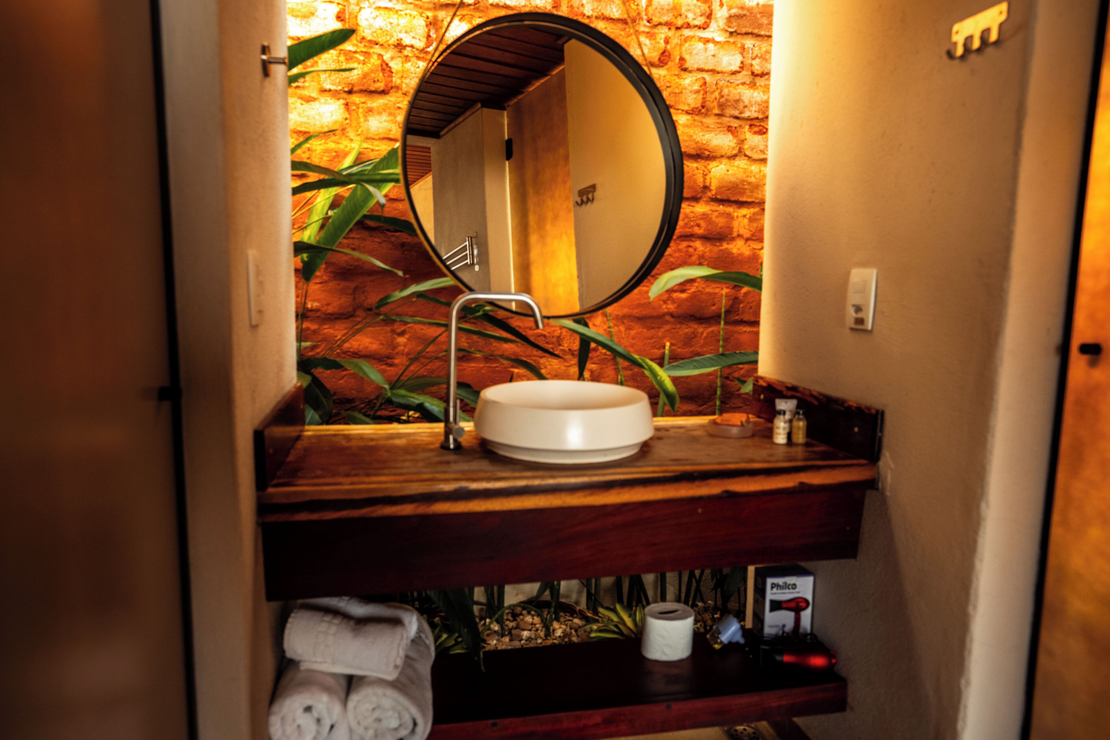 Baño con tocador de madera, espejo redondo y lavamanos. Hay toallas y artículos de aseo en el estante debajo del tocador.