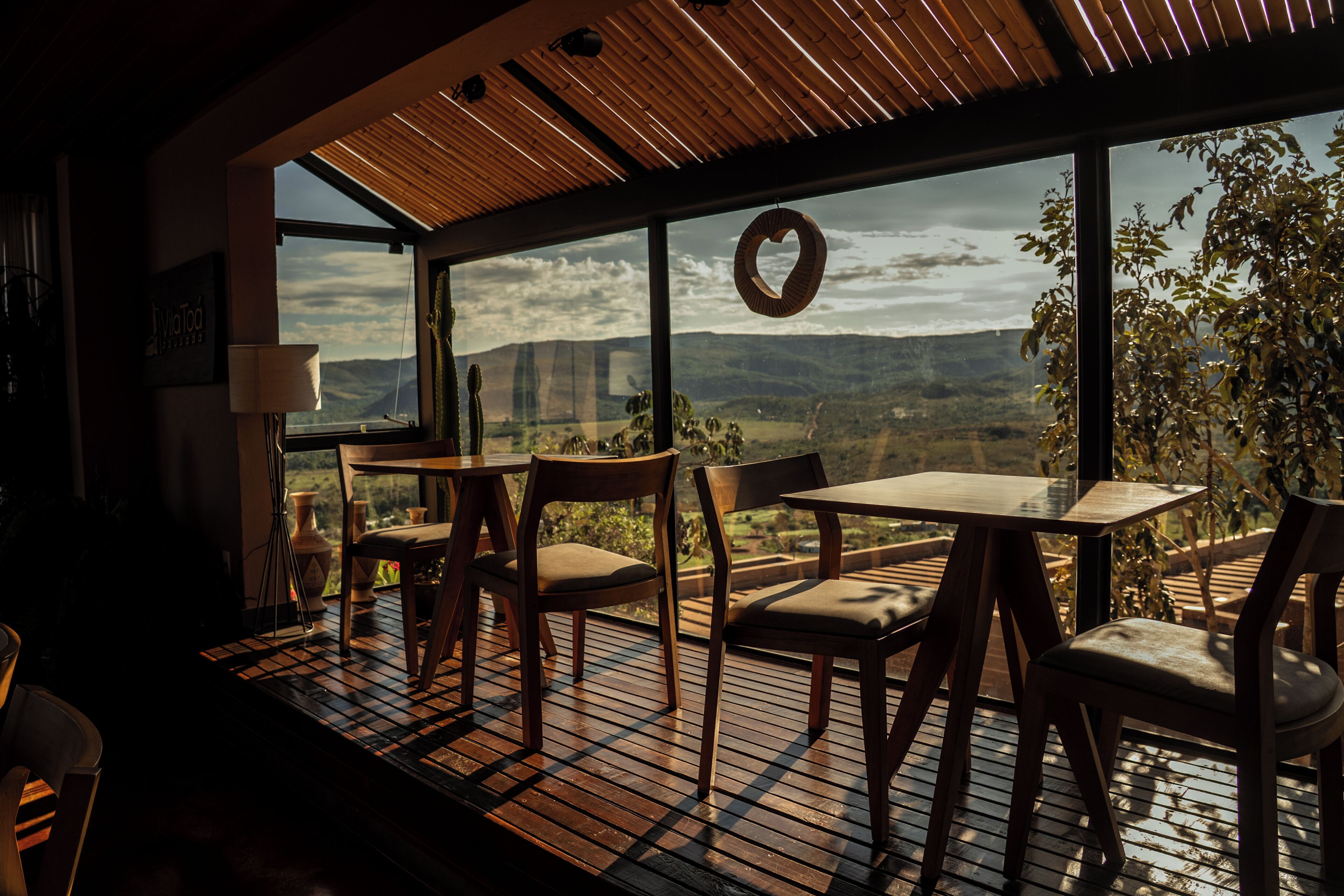 A dining area with a view of the countryside, featuring a table and chairs, a hanging wreath, and a wooden ceiling.