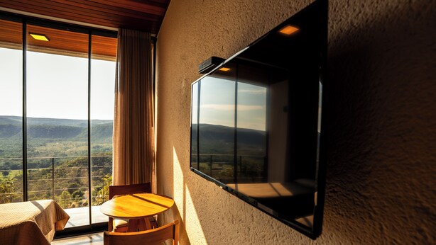 Una habitación con un ventanal grande, un televisor de pantalla plana montado en la pared, una mesa de madera y una silla.