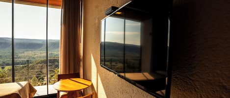 A room with a large window, a flat-screen TV mounted on the wall, a wooden table, and a chair.