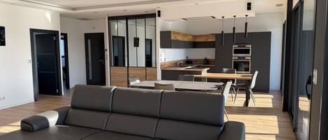 A modern living room with a grey sofa, a dining area with a table and chairs, and a kitchen with dark cabinets and a wooden countertop.