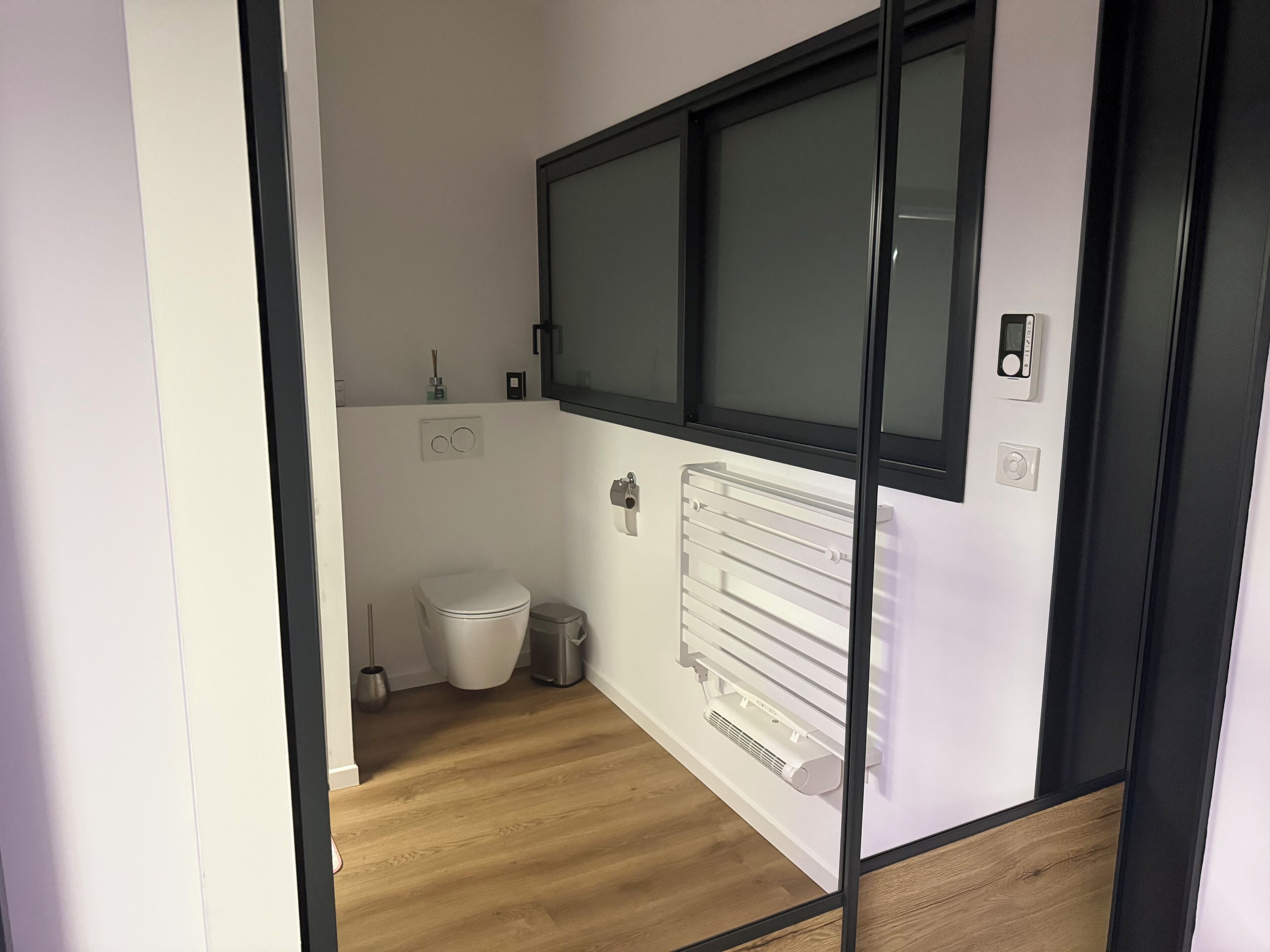 A small, modern bathroom with a toilet, a trash bin, a radiator, and a window.