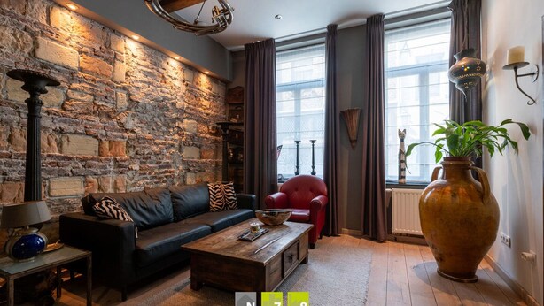 Ein gemütliches Wohnzimmer mit Steinwand, einer Ledersofa, einem roten Sessel und einer großen Vase mit Pflanze.