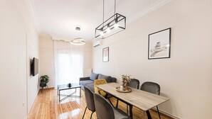 Ein modernes Wohnzimmer mit Holzboden, einem grauen Sofa, einem Esstisch mit Stühlen, einem an der Wand montierten Fernseher und einem gerahmten Bild.