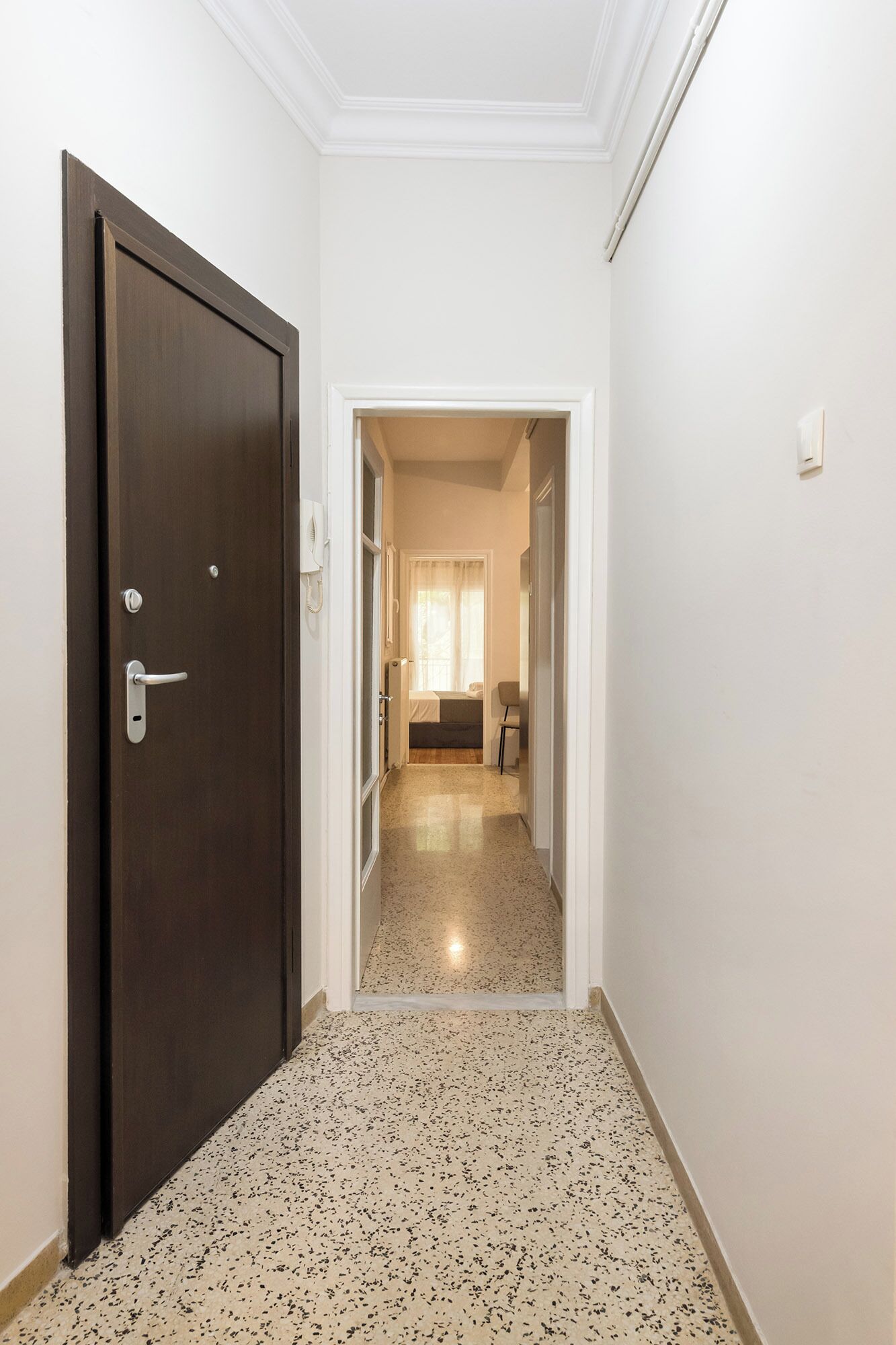 A hallway with a dark wooden door, a white wall, and a speckled floor.